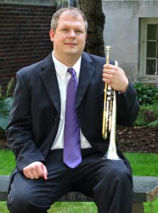 Adjunct faculty member, Keith Geiman sitting on bench outside holding a trumpet.